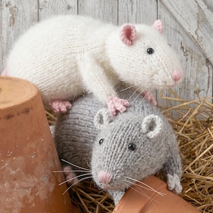May include: Two hand-knitted plush rats, one white and one gray, featuring black bead eyes, pink stitched noses, and long white whiskers. The white rat rests on top of the gray rat, both positioned near terracotta pots and scattered straw.