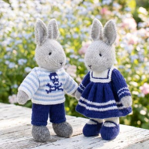 Könnte beinhalten: Zwei gestrickte Kaninchenpuppen mit grauem Fell. Das linke Kaninchen trägt einen hellblau-weiß gestreiften Pullover mit einem dunkelblauen Totenkopfmotiv und eine dunkelblaue Hose. Das rechte Kaninchen trägt ein dunkelblaues Kleid mit weißen Streifen und passenden Schuhen.