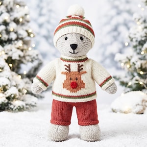 May include: A knitted bear toy wearing a cream-coloured sweater with a brown reindeer graphic featuring a red nose, red trousers, and a matching cream, red, and green striped knit hat with a pom-pom. The bear has light grey fur and black eyes.