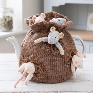 Pode incluir: Uma capa de malha castanha para bule com várias figuras pequenas de ratos de peluche presas ao exterior. Os ratos são de tons creme, cinzento-claro e branco, com orelhas e narizes cor-de-rosa. A capa tem uma textura canelada e topo franzido.