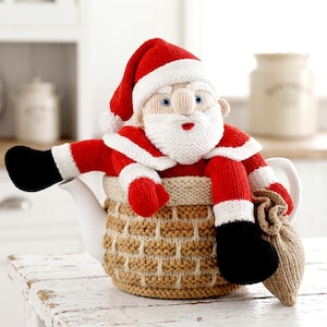 May include: A knitted tea cozy featuring a Santa Claus figure with a red hat, white beard, and red suit, sitting atop a tan basket-weave patterned base. The figure holds a small tan sack, and the entire piece is designed to fit over a white teapot.