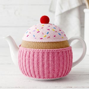 May include: A white ceramic teapot covered in a hand-knitted cosy designed to look like a cupcake. The cosy features a ribbed pink base, a tan middle section, and a light pink lid topper with colourful embroidered sprinkles and a red pom-pom.