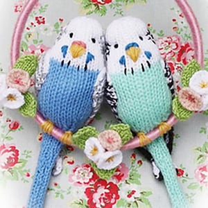 May include: Two knitted blue and green parakeets with black eyes and beaks are perched on a pink branch with white and pink flowers.