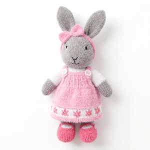 May include: A knitted grey rabbit doll wearing a pink dress with white accents and a floral pattern at the hem. The rabbit has a pink headband with a bow, white long sleeves, and matching pink shoes. The doll features embroidered facial details.