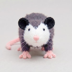 Op de afbeelding: Een klein, pluizig, gebreid opossum-figuurtje met een donkergrijs lichaam, een wit gezicht, zwarte oren, een lichtroze neus en lichtroze voetjes. Het wezen heeft lange, dunne witte snorharen en een lange, dunne roze staart aan de linkerkant.