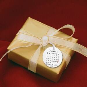Peut inclure: Une &eacute;tiquette cadeau ronde blanche avec un calendrier pour f&eacute;vrier 2075, avec la date du 24e encercl&eacute;e en rouge. L'&eacute;tiquette est attach&eacute;e &agrave; une bo&icirc;te cadeau dor&eacute;e avec un ruban blanc. Le texte sur l'&eacute;tiquette indique "F&eacute;vrier SMTWTFS 1 2 3 4 5 6 7 8 9 10 11 12 13 14 15 16 17 18 19 20 21 22 23 24 25 26 27 28 Sarah & Alex 2075"