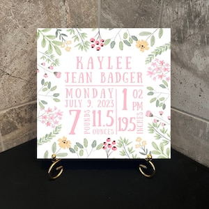 May include: A white ceramic tile with a floral design in pink, green, and yellow. The tile features the name "Kaylee Jean Badger", the date "Monday July 9, 2023", the time "1:02 PM", and the baby's birth stats: 7 pounds, 11.5 ounces, and 19.5 inches.