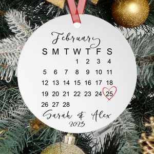 Peut inclure: Ornement rond blanc avec un calendrier pour f&eacute;vrier 2023, le 25 &eacute;tant encercl&eacute; et un c&oelig;ur rouge dessin&eacute; dessus. Le texte "Sarah & Alex" est &eacute;crit sous le calendrier.