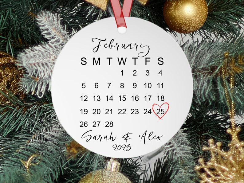 Peut inclure: Ornement rond blanc avec un calendrier pour f&eacute;vrier 2023, le 25 &eacute;tant encercl&eacute; et un c&oelig;ur rouge dessin&eacute; dessus. Le texte "Sarah & Alex" est &eacute;crit sous le calendrier.