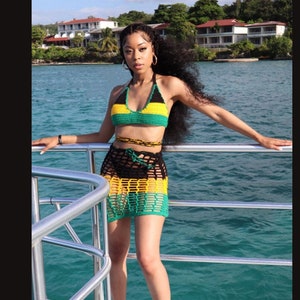 May include: A crochet bikini set in the colors of the Jamaican flag: black, yellow, and green. The set includes a halter top, a wrap-around waist tie, and a mini skirt. The woman is standing on a boat with the ocean in the background.