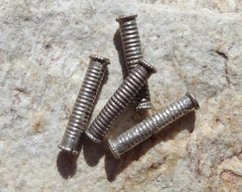 Könnte beinhalten: Eine Ansammlung kleiner, silberfarbener Metallperlen mit geripptem Design. Die zylindrischen Perlen haben dekorative Enden. Sie liegen auf einer hellen, strukturierten Steinoberfläche verstreut.