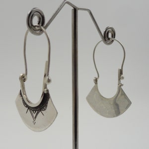 May include: A pair of silver hoop earrings with a geometric design. The earrings are hanging on a display stand.