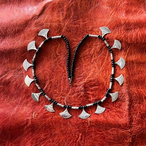May include: A silver necklace with black beads and silver crescent moon pendants. The necklace is laid out on a red leather surface.