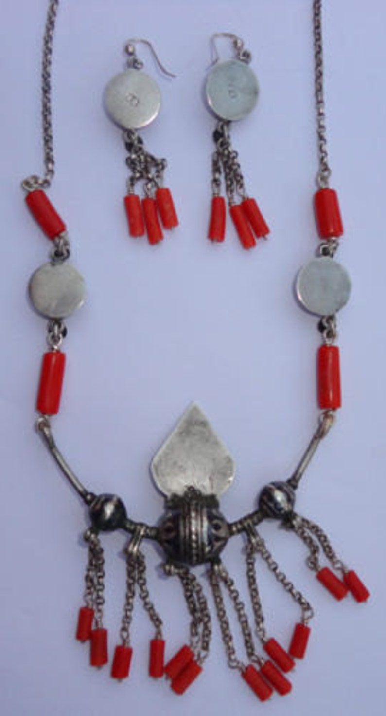 K&ouml;nnte beinhalten: Eine silberne Halskette mit roten Korallenperlen und einem silbernen Anh&auml;nger in Tropfenform. Die Halskette hat eine silberne Kette mit einem silbernen Verschluss. Der Anh&auml;nger hat ein silbernes Design in Tropfenform. Die Halskette wird mit einem Paar silberner Ohrringe mit roten Korallenperlen geliefert.