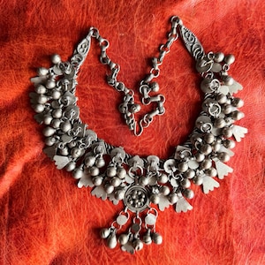May include: A silver-toned metal necklace featuring multiple dangling spherical bells and flat, fan-shaped charms. The piece includes a central circular medallion with a cluster of small spheres, connected by a chain with a hook-and-eye closure.