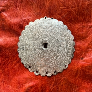May include: A silver circular pendant with a detailed, intricate design. The pendant has a scalloped edge and a small hole in the center.