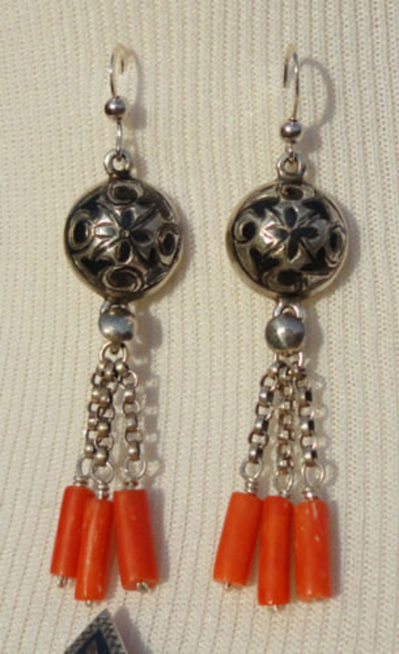 K&ouml;nnte beinhalten: Silberne Ohrringe mit einem schwarzen und silbernen Blumendesign und drei orangefarbenen Korallenperlen, die an jedem Ohrring h&auml;ngen.