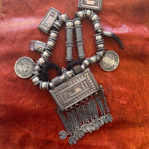 Puede incluir: Un collar de plata con un colgante rectangular, cuentas y dijes. El colgante tiene un diseño geométrico y está unido a una cadena con borlas. El collar también tiene un pequeño dije de cuerno de plata.