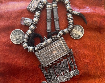 May include: A silver necklace with a rectangular pendant, beads, and charms. The pendant has a geometric design and is attached to a chain with tassels. The necklace also features a small silver horn charm.
