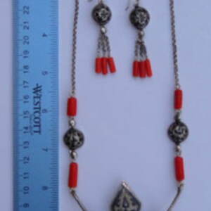 K&ouml;nnte beinhalten: Eine silberne Halskette mit einem Anh&auml;nger und roten Korallenperlen. Der Anh&auml;nger hat ein filigranes Silberdesign und ist an einer Kette mit mehreren Str&auml;ngen befestigt. Die Halskette wird mit passenden Ohrringen mit roten Korallenperlen geliefert.