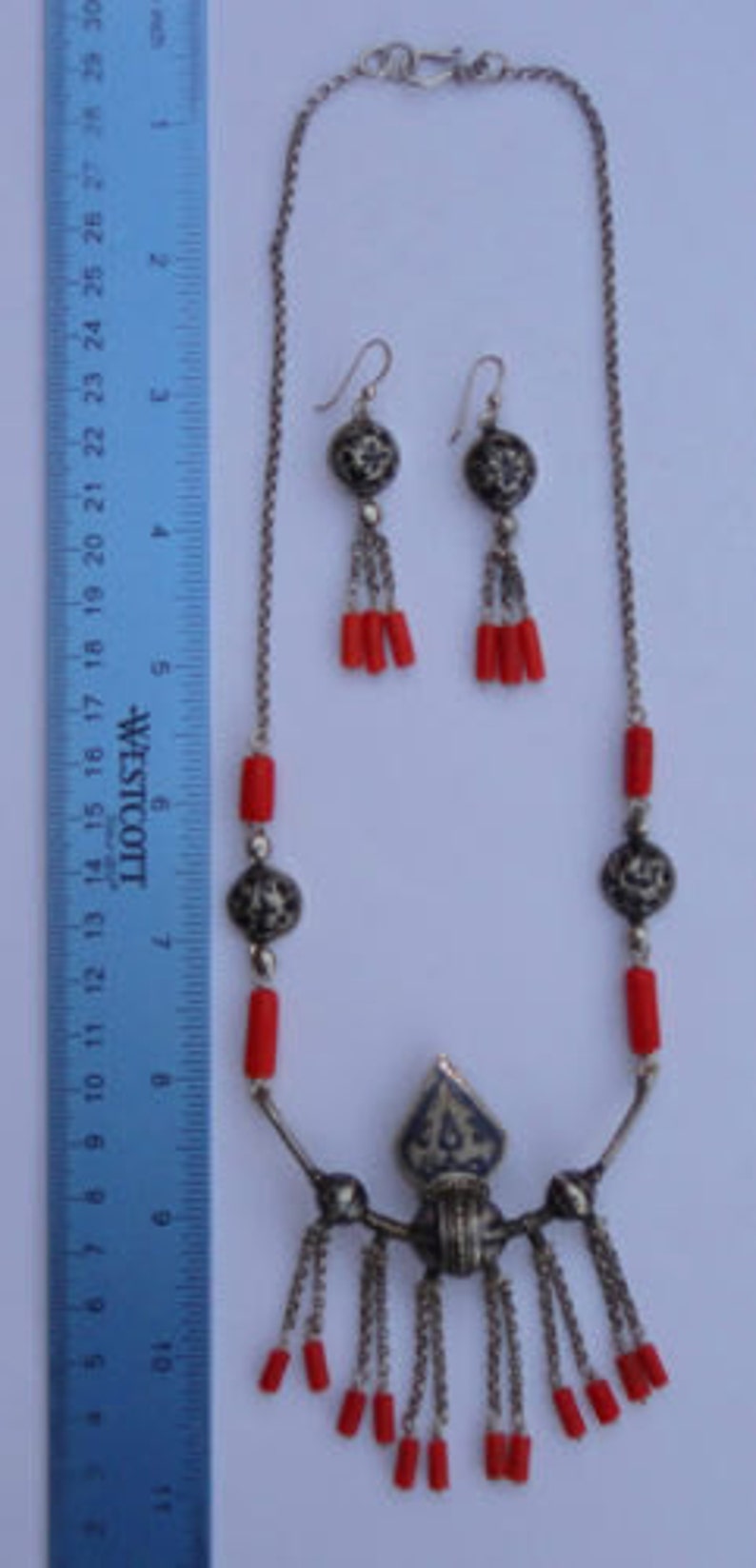 K&ouml;nnte beinhalten: Eine silberne Halskette mit einem Anh&auml;nger und roten Korallenperlen. Der Anh&auml;nger hat ein filigranes Silberdesign und ist an einer Kette mit mehreren Str&auml;ngen befestigt. Die Halskette wird mit passenden Ohrringen mit roten Korallenperlen geliefert.