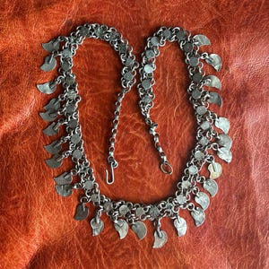 Può includere: Una collana a catena in metallo color argento caratterizzata da una serie di piccoli ciondoli piatti a forma di mezzaluna appesi a maglie circolari. La collana include una chiusura a gancio e una breve catenella di estensione.