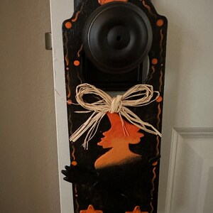 May include: A black Halloween door hanger with an orange witch silhouette and star accents. The door hanger is decorated with a raffia bow and orange polka dots. The door knob is black.