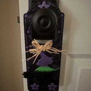 May include: Black Halloween door hanger with a witch silhouette in purple and green. The door hanger is decorated with purple stars and a raffia bow. The door knob is black.