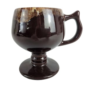 May include: A brown ceramic mug with a footed base and a brown and white glaze. The mug has a handle and is perfect for serving coffee or tea.