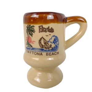 May include: A brown and beige ceramic mug with a handle. The mug has a design of a palm tree, a sailboat, and a wave with the text "Florida Daytona Beach".