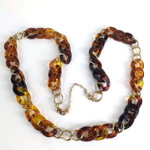 May include: A vintage necklace featuring large, oval-shaped links in shades of amber, brown, and yellow. The chain is connected with gold-tone metal rings and a delicate chain for closure. The necklace is a statement piece.