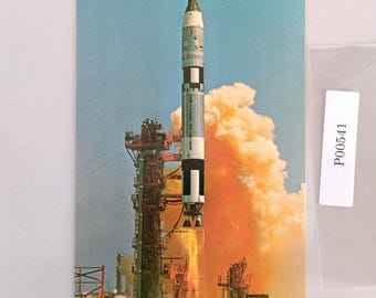 May include: Vintage postcard depicting a rocket launching into a blue sky, with flames and smoke billowing. The rocket is white, black, and silver, with "UNITED STATES" printed on the side. The postcard has a retro aesthetic.