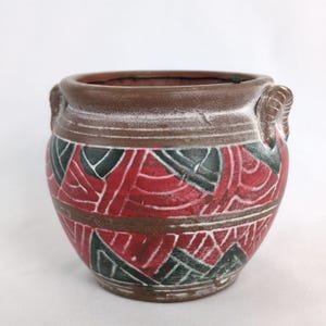 May include: A small, brown ceramic planter with two handles. The planter is decorated with a geometric pattern in red, green, and white.
