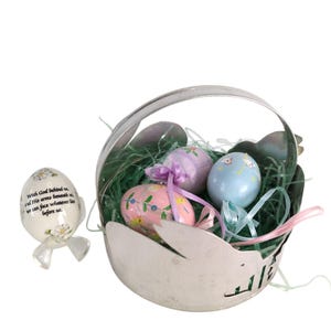 May include: A silver Easter basket filled with decorative eggs and green filler. The eggs are pastel colors with floral designs and ribbons. An egg with text and a bow is to the left. The text reads: "With God behind us, and His arms beneath us, we can face whatever lies before us."