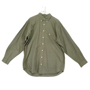 May include: An olive green long-sleeve button-down shirt featuring a pointed collar with buttons, a front placket with white buttons, and a single chest pocket on the left side with a small embroidered bird logo above the pocket opening.