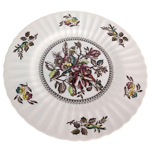 May include: White ceramic plate with a fluted edge and a floral design. The center features a bouquet of burgundy, teal, and brown flowers. Small floral and bird motifs are scattered around the rim.