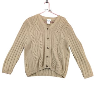 May include: Tan cable-knit cardigan sweater with a V-neck and button-up front. The long-sleeved sweater is hanging on a pink hanger. The sweater has a ribbed hem and cuffs.