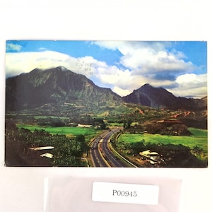 May include: A vintage postcard featuring a scenic landscape with mountains, a highway, and lush green fields under a cloudy blue sky. The postcard has the text "P00945" on a white label.