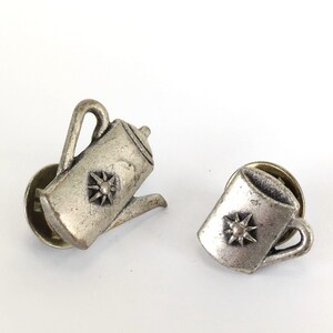 May include: Two silver-toned metal pins. One pin is a coffee pot shape, and the other is a mug shape. Both pins have a starburst design on the front. The pins have a classic, vintage aesthetic.