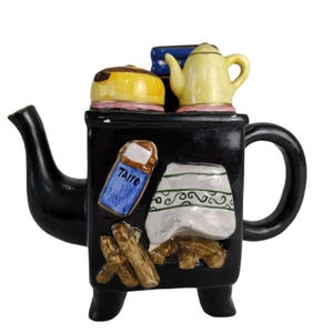 May include: Black ceramic teapot shaped like a wood-burning stove with a yellow teapot on top, a blue box with the word "Taiyo" on it, and a white cloth with green trim. The teapot has a black handle and spout.