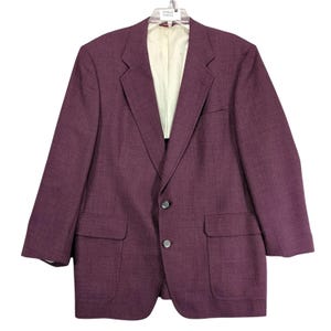 May include: A burgundy blazer with two front pockets and two-button closure. The blazer has a notched lapel and a light-colored lining. The garment is hanging on a hanger against a white background.