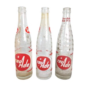 May include: Three vintage glass bottles of Tru-Ade, a carbonated beverage. The bottles are clear and have a red and white label with the brand name and a star.