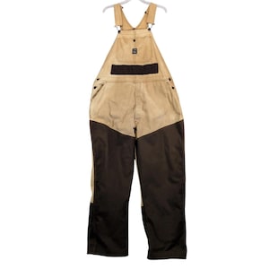 May include: Tan and dark brown overalls featuring a bib pocket with a small rectangular patch, adjustable shoulder straps with metal hardware, side buttons, and reinforced dark brown fabric panels covering the lower portion of the pant legs.