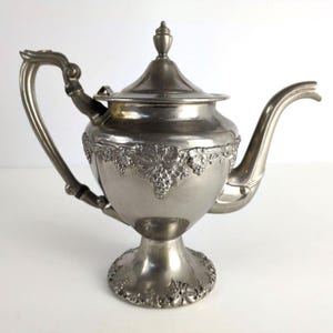 May include: Silver-colored teapot with a decorative grape and leaf design. The teapot has a curved handle, a spout, and a lid with a small finial. The base of the teapot is raised and has a detailed floral pattern.