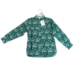May include: A long-sleeved button-down shirt in a deep forest green color featuring a repeating white pattern of evergreen trees and small snowflakes. The shirt includes a classic collar, a front button placket, and a white paper retail tag attached.