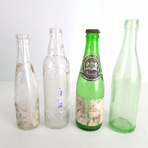 May include: Four vintage glass bottles of varying shapes and sizes. One is green with a label that reads "Serve Like Champagne Originals". The bottles are clear, with some showing signs of age and wear.