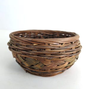 May include: A small, round woven basket made of brown and green natural materials. The basket has a sturdy, open weave design with a slightly flared shape. The rim is reinforced with tightly wound material, and the basket is suitable for storage or display.