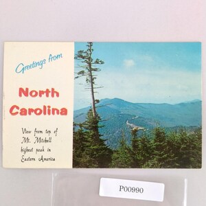 May include: Vintage postcard with a scenic view from the top of Mt. Mitchell in North Carolina. The postcard features a mountain range under a blue sky, with text that reads "Greetings from North Carolina".