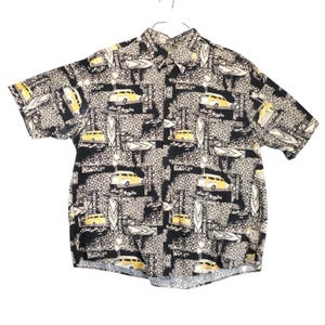 May include: Short-sleeved button-down shirt with a black, beige, and yellow pattern. The design features yellow cars, surfboards, and geometric shapes. The shirt is hanging on a white background.