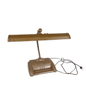 May include: A vintage desk lamp with a rectangular shade and a flexible gooseneck arm. The lamp is a muted gold color, with red and black buttons on top. The base has a handle and vertical lines. A black power cord extends from the base.